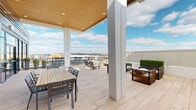 Located at 5801 Sanderson St NW in Huntsville, Alabama, this sophisticated rooftop terrace offers an exceptional outdoor amenity space designed for both relaxation and social gatherings. The terrace seamlessly blends covered and open-air zones, creating a versatile environment suited to a variety of uses throughout the year.

The covered portion features a striking warm wood-paneled ceiling with recessed lighting, supported by clean white structural columns that frame sweeping views of the Huntsville skyline and the rolling hills beyond. Large-format light-toned tile flooring extends across the entire terrace, lending a refined, cohesive aesthetic throughout.

The dining area is anchored by a slatted wood table paired with contemporary grey chairs, positioned to take advantage of natural light and the surrounding views. Floor-to-ceiling dark-framed glass doors and windows connect the terrace to an interior space, allowing abundant light to flow freely between the two.

The open-air section of the terrace is thoughtfully appointed with multiple seating arrangements, including a plush lounge set with neutral cushions and warm accent pillows, sleek chaise lounges, and modern accent furniture. Manicured boxwood hedges and tall planters add a touch of greenery and definition to the space.

Panoramic views of the developing Huntsville cityscape and the distant mountain ridgeline complete this elevated outdoor retreat.