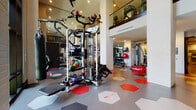 This well-appointed two-story fitness center offers a comprehensive workout environment designed with both function and aesthetics in mind. The main floor is expansive, featuring a dedicated cardio zone lined with treadmills and elliptical machines, including Cybex equipment, set against white-painted brick walls and accented by framed artwork. A strength training area houses a multi-functional cable machine, free weights, medicine balls, resistance equipment, and a weight bench, complemented by floor-to-ceiling mirrors and a large mounted flat-screen television. The space is unified by a bold hexagonal tile floor in a striking red, gray, and white pattern that adds visual energy throughout. White-painted brick columns punctuate the open floor plan, and ceiling fans ensure comfortable airflow. A motivational wall graphic reading "Don't Wish For It, Work For It" sets the tone of the space. The upper mezzanine level, accessible via an interior corridor with polished concrete floors, overlooks the main gym floor through a glass railing and wood-capped banister, and benefits from abundant natural light streaming through large steel-framed windows. This level provides additional equipment and a bird's-eye perspective of the full facility below.