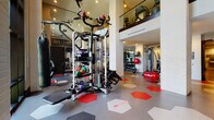This well-appointed two-story fitness center offers a comprehensive workout environment designed with both function and aesthetics in mind. The main floor is expansive, featuring a dedicated cardio zone lined with treadmills and elliptical machines, including Cybex equipment, set against white-painted brick walls and accented by framed artwork. A strength training area houses a multi-functional cable machine, free weights, medicine balls, resistance equipment, and a weight bench, complemented by floor-to-ceiling mirrors and a large mounted flat-screen television. The space is unified by a bold hexagonal tile floor in a striking red, gray, and white pattern that adds visual energy throughout. White-painted brick columns punctuate the open floor plan, and ceiling fans ensure comfortable airflow. A motivational wall graphic reading "Don't Wish For It, Work For It" sets the tone of the space. The upper mezzanine level, accessible via an interior corridor with polished concrete floors, overlooks the main gym floor through a glass railing and wood-capped banister, and benefits from abundant natural light streaming through large steel-framed windows. This level provides additional equipment and a bird's-eye perspective of the full facility below.