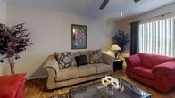 This single-floor residence offers a thoughtfully organized layout with multiple living spaces connected by warm wood-style flooring that runs throughout the home. The living room serves as a central gathering area, featuring a textured ceiling, a ceiling fan, and vertical blinds that filter natural light through a large window. The space accommodates ample seating and opens to a defined dining area, which includes a glass-top table set beneath overhead lighting and is accented by decorative wall art.

The kitchen is efficiently designed with white cabinetry, light countertops, a stainless steel sink, and black appliances including a dishwasher. Open shelving above provides additional storage and display space.

The home includes multiple bedrooms, each equipped with ceiling fans, wood-style flooring, and vertical blinds or curtained windows. One bedroom features a sliding glass door that opens to an outdoor view, providing a connection to the surrounding greenery. Both bathrooms are finished with white vanities, large mirrors, and tub-shower combinations with decorative curtains, offering functional and clean spaces throughout.