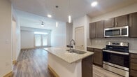 This well-appointed 1-bedroom, 1-bathroom residence is thoughtfully designed with modern finishes and an efficient single-floor layout. The open-concept kitchen serves as the heart of the home, anchored by a large granite-topped island with an undermount sink and elegant pendant lighting. Dark espresso cabinetry with brushed hardware contrasts beautifully against the light granite countertops and classic subway tile backsplash. A full suite of stainless steel appliances, including a refrigerator, range, microwave, and dishwasher, completes the kitchen. The kitchen flows seamlessly into the living area, where wood-look plank flooring, a ceiling fan, and a set of glass-paned double doors invite natural light and create a bright, airy atmosphere. The bedroom offers a comfortable retreat with plush carpeting, dual windows with blinds, a ceiling fan, and ample wall space. The bathroom is finished with the same cohesive design language, featuring a dark wood vanity with granite countertop, an undermount sink, a tiled shower, and light flooring. Recessed lighting and clean white walls throughout the residence contribute to a fresh, contemporary ambiance.