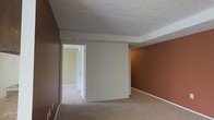 This single-floor residential space offers a straightforward and functional layout designed for comfortable everyday living. The interior is characterized by a warm, earthy color palette, with rich terracotta-toned accent walls contrasting against softer cream and white surfaces to create a grounded, inviting atmosphere throughout.

The main living area features wall-to-wall carpeting in a neutral beige tone, contributing to a soft underfoot feel and a cohesive aesthetic. The textured popcorn ceiling adds a classic residential finish overhead, while white trimwork and baseboards provide clean visual boundaries throughout the space.

An open doorway connects the main room to an adjacent hallway or secondary room, suggesting a practical flow between spaces. The connecting area benefits from lighter wall tones, offering a subtle contrast to the deeper hues of the primary room. A white paneled door is visible in the background, adding to the traditional residential character of the space.

Overall, the space presents a warm and unpretentious interior, well-suited for those seeking a comfortable, move-in-ready environment. The consistent use of carpet flooring, neutral trim, and bold accent walls gives the space a defined personality while maintaining versatility for personalization.