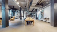 This expansive single-floor space masterfully blends industrial heritage with curated character. Soaring ceilings reveal exposed ductwork, conduit runs, and track lighting mounted on steel beams, while polished and terrazzo concrete floors stretch throughout, lending the interior a raw yet refined quality. Structural columns in dark steel punctuate the open floor plan, reinforcing the building's industrial origins.

The main area is defined by a large communal wood farm table surrounded by metal chairs, offering a central gathering point. Original brick walls and large black-framed steel windows and doors allow natural light to filter in, complementing the eclectic mix of vintage and contemporary furnishings — including wingback chairs, chesterfield sofas, and accent ottomans. A standout antique wooden apothecary cabinet with brass hardware serves as both a functional and decorative focal point alongside gallery-style artwork displayed throughout.

A reception or front desk area anchors one end of the space, accompanied by high-top tables and stools. Mailbox installations and directional signage suggest a multi-use or shared-occupancy building. The varied zones — from collaborative work areas to lounge nooks — create a dynamic, layered environment rich in texture and creative energy.