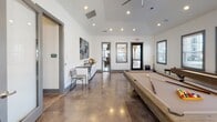 This single-floor amenity space is thoughtfully designed to serve as a versatile hub for recreation, work, and socializing. The open layout is anchored by polished concrete floors that reflect the abundant natural light streaming in through multiple large windows fitted with blinds, giving the space a clean, contemporary feel.

At the heart of the room sits a substantial pool table with a rustic wood frame, complemented by a full-size shuffleboard table along one wall — making this an ideal gathering spot for leisure and entertainment. A ceiling fan overhead ensures comfort throughout.

Along one wall, a dedicated workstation area features multiple computer desks with ergonomic chairs, ideal for productivity needs. Above the workstations, a striking collection of organic wood and sculptural wall art adds an artistic focal point. A small bistro table nearby offers an informal spot to sit and unwind.

On the opposite side of the room, a built-in wet bar with white cabinetry, a dark countertop, and a sink provides convenient refreshment access. Bold abstract artwork in blue and gold tones adorns the wall above, adding a vibrant splash of color.

The vaulted ceiling with recessed lighting enhances the sense of openness throughout, while glass-panel doors connect the space to adjacent areas and the outdoors, creating a bright and welcoming atmosphere.
