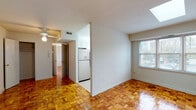 This single-floor unit offers a well-organized layout that makes efficient use of its space. The most distinctive design feature found throughout the living and sleeping areas is the richly toned parquet hardwood flooring, which adds warmth and a timeless character to every room. A skylight is a notable architectural highlight, allowing natural light to pour into the main living area, complementing the large windows that frame outdoor views of surrounding greenery.

The living area is open and versatile, with neutral white walls that provide a clean backdrop. A ceiling fan overhead adds both function and a casual aesthetic to the space. The kitchen is tucked neatly off the main living area and is equipped with stainless steel appliances, including an electric range with an over-the-range microwave, a full-size refrigerator, and white cabinetry paired with light countertops, creating a bright and practical cooking space.

The bathroom is finished in crisp white throughout, featuring a full bathtub with shower, a white vanity with under-sink storage, mosaic tile flooring, and a frosted window that introduces natural light while maintaining privacy. The bedroom is a quiet, well-lit room with the same parquet flooring and clean white walls, offering a calm and restful environment. Overall, the unit presents a cohesive, light-filled living space with classic finishes.