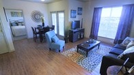 This single-floor residence features an open-concept layout that seamlessly integrates the living and dining areas into one cohesive, functional space. The hardwood-style flooring extends throughout, providing a warm and consistent visual flow from one zone to the next.

The living area is anchored by a rectangular coffee table set atop a colorful, polka-dot area rug, complemented by a light blue accent chair that adds a pop of color. A dark media console serves as the entertainment focal point, topped with a flat-screen television and flanked by decorative artwork on the walls. A large window dressed in deep purple curtains allows natural light to fill the space while adding a bold design element.

The adjacent dining area features a dark wood high-top table with matching bar-height chairs, positioned near a set of glass patio doors that invite ample natural light and provide outdoor access. A decorative sunburst mirror and fresh floral arrangement add character to the dining wall.

Greenery in the form of potted plants adds life and texture throughout, and the overall ambiance strikes a balance between casual comfort and thoughtful, contemporary styling.