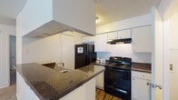 Located at 8730 N Himes Ave in Tampa, Florida, this single-floor unit offers a practical and well-organized layout. The kitchen is a standout feature, equipped with dark granite-style countertops, white cabinetry, and a full suite of black appliances including a refrigerator, electric range, and built-in range hood. A peninsula counter provides additional workspace and a natural transition into the adjacent living area. The flooring in the kitchen is a warm wood-look laminate, adding contrast to the bright white cabinetry. The living area is spacious and open, featuring neutral wall-to-wall carpeting, white walls, and a textured ceiling. Natural light filters in through a window fitted with blinds, and a front entry door with a modern keypad lock provides secure access. The bedroom continues the carpeted flooring and offers ample space, with a large closet fitted with vertical blinds and a window that brings in additional natural light. The bathroom includes a tub-shower combination, a toilet, and a vanity with dark countertop and white cabinetry, complemented by overhead storage and a mirrored medicine cabinet. Wood-look flooring in the bathroom ties back to the kitchen's aesthetic, giving the unit a cohesive feel throughout.