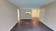 This single-floor residence is part of a well-kept multi-building community characterized by lush landscaping, mature trees, manicured gardens with colorful flower beds, and a gated parking area with covered carports. The grounds are thoughtfully designed with paved walkways, decorative rock arrangements, and abundant greenery that contribute to a welcoming, park-like atmosphere. The exterior of the buildings features a classic two-tone design with wood-accented balconies, giving the community a warm, established aesthetic.

Inside, the open-concept living and dining area is finished with rich, dark hardwood-style flooring that runs throughout, offering a clean and modern look. Neutral-toned walls complement the light-colored baseboards and trim, creating a bright and airy ambiance. A textured ceiling adds character to the space. The kitchen connects to the main living area via a pass-through window, enhancing the open flow between spaces. The dining area is equipped with a ceiling fan, providing both functionality and comfort. Natural light filters into the interior, contributing to a welcoming and comfortable atmosphere throughout the residence.
