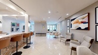 Located at 1900 Yorktown Street in Houston's Galleria area, this thoughtfully designed single-floor communal space offers a generous and versatile layout across two connected living areas. Polished marble-look tile flooring flows seamlessly throughout, reflecting natural light that pours in through large windows framing views of lush greenery outside.

The first living area serves as a welcoming lounge, anchored by textured architectural columns clad in woven wall covering. This zone features curated seating arrangements, pendant lighting with sleek black fixtures, and carefully selected artwork that adds warmth and character. A built-in bar counter with rich stone cladding and barstool seating provides an ideal gathering point, complemented by a wall-mounted television nearby.

The second living area is a more expansive, elegantly appointed lounge featuring a bold blue-toned area rug, a mix of plush sofas and accent chairs, and recessed lighting overhead. Multiple windows dressed with roller shades fill the room with soft, diffused natural light while maintaining privacy. Curated contemporary artwork adorns the white walls throughout, enhancing the modern aesthetic.

Together, the two living areas create a cohesive and inviting environment that balances functionality with sophisticated design, making this space well-suited for both relaxed socializing and more structured gatherings.