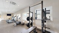 This fitness center in Longview, Texas offers a comprehensive workout facility within a single, open-plan space. The layout is thoughtfully organized to accommodate both cardio and strength training needs. Along one side, a row of commercial-grade treadmills, elliptical machines, and a stationary bike are positioned to take advantage of the large windows that line the walls, offering natural light and views of the surrounding outdoor pool area and community grounds. A wall-mounted flat-screen television provides entertainment for cardio sessions. On the strength training side, a Rogue power rack is fully loaded with barbells and an extensive selection of weight plates, accompanied by a flat utility bench. A dumbbell rack with a full range of free weights and exercise accessories such as a stability ball and step platform round out the offering. Large wall mirrors in the strength area enhance the sense of space and allow for form monitoring during lifts. The flooring throughout is durable rubber-style material suited for heavy use, while the drop ceiling with recessed lighting ensures bright, even illumination. The space is accessed through elegant French doors, adding a refined touch to this practical and well-appointed fitness facility.
