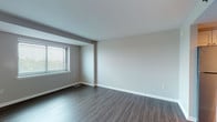 This single-floor residence offers a thoughtfully designed layout that balances open communal areas with private living spaces. The main living and dining area is expansive and airy, featuring dark hardwood-style plank flooring, neutral gray walls, and wide windows fitted with horizontal blinds that allow generous natural light to filter in. Structural columns and ceiling beams add architectural character to the open-plan space.

The kitchen is well-equipped with white cabinetry, dark granite countertops, a stainless steel refrigerator, a dishwasher, and recessed lighting. It opens toward the front entryway, which features tile flooring and a paneled white door.

The two bedrooms are carpeted in warm neutral tones and each benefit from broad windows that bring in ample daylight and outdoor views. The primary bathroom is bright and spacious, with white tile flooring, a white vanity topped with dark granite, a large mirror, and a soaking tub. A second bathroom features a sliding glass shower enclosure, a matching white vanity with granite countertop, and grab bars for accessibility.

A dedicated laundry area houses a front-loading washer and a wall-mounted drying rack, offering added everyday convenience. The overall aesthetic is clean, modern, and move-in ready.