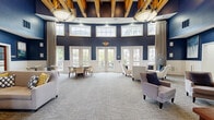 Located in Annapolis, MD, this well-appointed clubhouse offers a sophisticated and welcoming environment designed for community gatherings and social events. The single-floor layout encompasses a spacious living room and a fully equipped kitchen, together delivering a refined yet functional space.

The living room is the centerpiece of the clubhouse, showcasing soaring ceilings adorned with exposed wood beam detailing and dramatic navy blue walls accented by white wainscoting. Towering natural stone columns flank a classic stone fireplace, lending a warm, lodge-inspired character to the room. Abundant natural light pours in through large French doors and upper clerestory windows, while elegant pendant light fixtures illuminate the space. Plush patterned carpeting and carefully arranged seating areas create an inviting atmosphere throughout.

The adjacent gathering area features a rounded design with floor-to-ceiling drapery, additional French doors opening to the outdoors, and stylish upholstered seating ideal for socializing.

The kitchen is well-resourced with rich cherry wood cabinetry, granite countertops, a central island with gas cooktop and stainless steel range hood, and high-end stainless steel appliances. Stone tile flooring and ample natural light from Roman shade-dressed windows complete the space. A casual dining area with bistro-style tables provides a relaxed spot for informal gatherings.