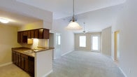 This single-floor residence offers a thoughtfully designed open-concept layout that seamlessly connects the kitchen and living areas. The kitchen is appointed with rich dark wood cabinetry, granite-look countertops, and a peninsula that provides both additional workspace and a natural divider between the cooking and living spaces. An elegant pendant light hangs above the peninsula, adding a warm focal point to the area.

The expansive living area is defined by soaring vaulted ceilings that amplify the sense of space and volume throughout. Plush wall-to-wall carpeting runs across the living space, contributing to a comfortable and welcoming ambiance. A ceiling fan is centrally positioned, offering both practical airflow and a subtle design element.

Natural light fills the space generously through multiple windows and glass-paneled doors along the far wall, creating a bright and airy atmosphere throughout the day. The neutral color palette of warm beige and white tones on the walls and trim enhances the openness of the interior. The layout offers a smooth, unobstructed flow from the kitchen through to the living area, making the space both functional and inviting for everyday living and entertaining.