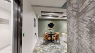 This thoughtfully designed single-floor amenity space combines a premium indoor golf simulator with an elegantly appointed lounge area, offering a refined recreational experience. The space is organized into distinct zones that flow seamlessly together.

The lounge area sets a sophisticated tone with sage-green accent walls, a decorative gold-framed organic mirror, and a bold coffered ceiling with recessed lighting. Lush, floral-patterned carpeting in muted greens, golds, and grays anchors the space, complementing the warm amber velvet dining chairs surrounding a dark round table. A separate seating nook features a curved white accent chair, a plush gray sofa, and a marble-topped side table with a stylish lamp, creating an inviting space for relaxation or socializing.

A dedicated tech station offers a wall-mounted screen flanked by sleek vertical sconce lighting and a floating white ledge with coordinating green ottomans beneath — ideal for reviewing game stats or casual viewing.

The simulator bay is dramatically enclosed with dark walls and blue ambient lighting, featuring a large projection screen and a bright green synthetic turf putting mat. A projector mounted overhead, golf clubs, and a compact storage unit complete this functional yet atmospheric zone. A textured botanical wallcovering on one wall adds visual interest and reinforces the space's upscale character.