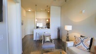 This single-floor residence offers a thoughtfully designed open-concept layout that seamlessly connects the kitchen, dining, and living areas into one cohesive living space. Rich, dark hardwood floors run throughout, grounding the interior with warmth and contrast against the neutral, light-toned walls that rise to impressive heights, lending a sense of spaciousness and airiness to the floor plan.

The kitchen is well-appointed with stainless steel appliances, including a refrigerator and microwave, complemented by light wood cabinetry and a granite or stone-topped breakfast bar. Pendant lighting hangs above the bar, providing both functional illumination and a decorative accent. Bar stools at the counter offer a casual dining option, making the kitchen an inviting hub for everyday living.

The living area is warm and comfortable, furnished with plush seating and accented by a statement floor lamp. The open sight lines between the kitchen and living spaces promote an easy flow, ideal for entertaining or relaxed daily life. Subtle decorative touches, including accent pillows and natural-toned decor elements, contribute to the space's refined yet approachable character. Overall, the residence presents a clean, contemporary aesthetic well-suited for modern urban living.