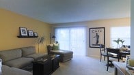This single-floor space centers around a well-appointed living area that balances comfort and style. The room is anchored by a large sliding glass door dressed with vertical blinds, allowing abundant natural light to fill the space while offering access to the outdoors. Warm yellow walls lend a welcoming, energetic ambiance to the interior, complementing the neutral carpeted flooring throughout.

The living area is furnished with a plush sofa and a chaise lounge accented with green throw pillows, creating an inviting seating arrangement. A dark wood coffee table serves as a functional centerpiece, grounding the layout. A floor lamp provides additional lighting options, while a wall-mounted shelf displays framed artwork and a decorative plant, adding personality to the space.

A framed café-style poster adorns one wall, contributing a cosmopolitan aesthetic, while a small dining area with a dark chair is partially visible near the corner. The textured ceiling and clean white trim offer a classic architectural finish. Overall, the space presents a cozy yet stylish environment with practical layout and ample natural light.