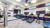 Located on Inspiration Ave in Bluffton, SC, this single-story fitness center offers a generously sized, thoughtfully designed space dedicated to health and exercise. The facility is divided into two primary workout zones, each distinguished by a bold interior design featuring alternating purple and white horizontal stripes across the walls, creating an energetic and motivating atmosphere throughout.

The cardio area is equipped with multiple treadmills and stationary bikes, all positioned facing a large flat-screen television mounted on a full-length mirrored wall. The mirrors effectively expand the sense of space and allow users to monitor their form during workouts. Multiple ceiling fans maintain airflow, while flush-mounted panel lighting ensures consistent illumination. Windows along the exterior wall allow natural light to filter in, offering views of surrounding greenery.

The strength training zone features a cable machine, a functional training rack with suspension straps, a punching bag, a bench press station, and a variety of free weights and medicine balls. Wall-mounted mirrors are present here as well, complementing the playful polka-dot accent wall detail in this area. Light-toned checkerboard carpet tile flooring unifies the space, providing both durability and comfort underfoot.