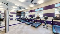 Located on Inspiration Ave in Bluffton, SC, this single-story fitness center offers a generously sized, thoughtfully designed space dedicated to health and exercise. The facility is divided into two primary workout zones, each distinguished by a bold interior design featuring alternating purple and white horizontal stripes across the walls, creating an energetic and motivating atmosphere throughout.

The cardio area is equipped with multiple treadmills and stationary bikes, all positioned facing a large flat-screen television mounted on a full-length mirrored wall. The mirrors effectively expand the sense of space and allow users to monitor their form during workouts. Multiple ceiling fans maintain airflow, while flush-mounted panel lighting ensures consistent illumination. Windows along the exterior wall allow natural light to filter in, offering views of surrounding greenery.

The strength training zone features a cable machine, a functional training rack with suspension straps, a punching bag, a bench press station, and a variety of free weights and medicine balls. Wall-mounted mirrors are present here as well, complementing the playful polka-dot accent wall detail in this area. Light-toned checkerboard carpet tile flooring unifies the space, providing both durability and comfort underfoot.