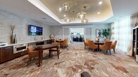 Located at 1055 Hatches Pond Lane in Cary, NC, this generously sized single-floor community space offers an impressive array of amenities finished with upscale materials and thoughtful design throughout. The layout flows seamlessly from one area to the next, beginning with a large, open dining area featuring large-format porcelain tile floors laid in a striking herringbone pattern, crisp white columns, tray ceilings with wood accents, and abundant natural light streaming through floor-to-ceiling arched windows. Multiple dining configurations accommodate both intimate and larger group settings. An adjacent dining room with patterned carpeting and tufted upholstered chairs provides a warmer, more intimate atmosphere, with direct access to an outdoor patio area. The professional-grade kitchen is a standout feature, centered around an oversized marble island with dramatic veining, complemented by rich dark wood cabinetry, stainless steel appliances, a herringbone tile backsplash, and elegant pendant lighting. The theater room offers tiered seating with plush swivel chairs, a large wall-mounted display, and a tray ceiling with ambient lighting. A library and study nook rounds out the space, featuring built-in bookshelves, a workstation, and curated artwork — creating a quiet retreat within this dynamic community hub.