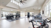 Located at 7301 Topwater Dr in Wilmington, NC, this expansive fitness center offers a generously sized, single-floor exercise facility designed to support a full range of workout routines. The space features impressively high ceilings in the main area, accentuated by large clerestory windows that flood the interior with natural light and oversized ceiling fans that promote airflow throughout.

The main exercise floor is finished with durable, easy-to-clean epoxy-style flooring and is organized into distinct training zones. The cardio section houses a full lineup of equipment including treadmills, ellipticals, stair climbers, stationary bikes, and a Peloton cycle. The strength training area offers cable machines, a multi-station rack with free weights, adjustable benches, and a comprehensive dumbbell rack displayed along a wall of mirrors for form feedback.

A dedicated functional training annex, accessible through elegant arched doorways, features exposed wood beam ceilings, stylish wicker pendant lighting, TRX suspension trainers, battle ropes, an air bike, medicine balls, and foldable floor mats — creating a boutique studio atmosphere. Large glass doors and windows throughout provide outdoor views of the surrounding green landscape. Tasteful wall art and textured accent wallpaper add a polished, welcoming aesthetic to the overall space.
