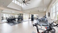 Located at 7301 Topwater Dr in Wilmington, NC, this expansive fitness center offers a generously sized, single-floor exercise facility designed to support a full range of workout routines. The space features impressively high ceilings in the main area, accentuated by large clerestory windows that flood the interior with natural light and oversized ceiling fans that promote airflow throughout.

The main exercise floor is finished with durable, easy-to-clean epoxy-style flooring and is organized into distinct training zones. The cardio section houses a full lineup of equipment including treadmills, ellipticals, stair climbers, stationary bikes, and a Peloton cycle. The strength training area offers cable machines, a multi-station rack with free weights, adjustable benches, and a comprehensive dumbbell rack displayed along a wall of mirrors for form feedback.

A dedicated functional training annex, accessible through elegant arched doorways, features exposed wood beam ceilings, stylish wicker pendant lighting, TRX suspension trainers, battle ropes, an air bike, medicine balls, and foldable floor mats — creating a boutique studio atmosphere. Large glass doors and windows throughout provide outdoor views of the surrounding green landscape. Tasteful wall art and textured accent wallpaper add a polished, welcoming aesthetic to the overall space.