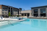 This outdoor amenity area in Meridian, Idaho, is a well-appointed resort-style pool and spa complex designed for relaxation and recreation. At its center sits a generously sized swimming pool with crystal-clear water, complemented by an integrated spa with tiered, wood-accented surround that adds a warm, contemporary touch to the overall design. The pool deck is finished in smooth concrete and extends broadly around the water, offering ample space for lounging and socializing. Rows of white chaise lounges and Adirondack-style chairs are arranged throughout the deck, with shaded umbrella groupings and accent tables providing comfortable spots for gathering. A dedicated outdoor ping-pong table adds a recreational element to the space. The area is enclosed by a sleek black metal fence, with landscaped planters and young trees providing greenery and visual interest along the perimeter. Adjacent to the pool, a modern building houses the spa and pool facilities, featuring floor-to-ceiling glass panels that connect the indoor and outdoor environments. Signage indicates designated spa and pool hours and guidelines. The surrounding multi-story buildings frame the courtyard setting, while the open sky above floods the entire area with abundant natural light, creating a bright, vibrant outdoor retreat.
