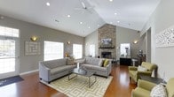 This single-story space makes a striking impression through its expansive living area, defined by soaring vaulted ceilings that amplify the sense of openness and volume. A ceiling fan is centrally mounted, complementing the recessed lighting embedded throughout the ceiling to provide layered illumination across the room.

The focal point of the living area is a floor-to-ceiling stacked stone fireplace, which anchors the space with a bold, textural presence and serves as a natural gathering point. Warm hardwood flooring spans the room, adding richness and continuity underfoot, while neutral gray walls create a clean, versatile backdrop.

Natural light enters generously through multiple large windows positioned along the walls, brightening the interior and enhancing its airy atmosphere. The living area is thoughtfully arranged with two gray tufted sofas oriented around a metal coffee table, set atop a geometric-patterned area rug in green and white tones.

Wall sconces add ambient warmth alongside the recessed lighting, contributing to a balanced and inviting atmosphere. The overall design blends casual comfort with architectural character, making this a functional yet visually engaging living environment.