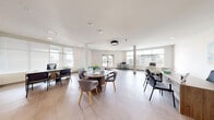 Located at 6834 Milwaukee St in Madison, Wisconsin, this single-floor office space offers a well-appointed, open-plan layout designed to accommodate a variety of work and meeting needs. The interior is finished with large-format neutral tile flooring throughout, white walls, and smooth ceilings with recessed lighting complemented by distinctive circular pendant fixtures, creating a bright and professional atmosphere.

The space is organized into several functional zones. A central round table with upholstered grey chairs serves as a collaborative hub, ideal for group discussions or informal meetings. Multiple individual workstations and desks are positioned around the perimeter, offering dedicated areas for focused work. A comfortable lounge corner features bold blue accent chairs alongside a vibrant abstract wall art piece, adding a creative touch to the environment. A teal modular seating unit near the entrance provides an additional casual gathering spot.

Large windows on multiple walls flood the space with natural light and offer views of the surrounding exterior. Structural columns are integrated throughout the open floor plan. Decorative tall grass planters in navy pots add a natural, organic element to the space. A printer station and additional worktables round out the practical amenities. Direct access to the exterior is provided via a glass-panel door, enhancing connectivity and ease of access.
