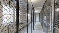 This storage room is organized around a central concrete-floored corridor lined on both sides with individual wire-mesh caged storage units. Each unit is constructed from sturdy steel chain-link fencing and framed with metal posts, reaching considerable heights to maximize vertical storage capacity. The units are secured with individual locks, providing dedicated, private storage for each user. The open mesh design of the caging allows visibility into each unit while maintaining clear boundaries between spaces. Natural light filters into the corridor through windows visible within some of the individual units, supplementing the overhead artificial lighting that illuminates the walkway. The perimeter walls are composed of painted concrete block, contributing to the utilitarian character of the space. Fire safety equipment, including alarm components, is visibly mounted along the walls, underscoring the room's compliance with safety standards. The corridor between the units is kept clear, allowing straightforward access to each caged space. Overall, the storage room presents a well-organized, functional environment designed to provide secure, individual storage in a shared setting.