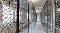 This storage room is organized around a central concrete-floored corridor lined on both sides with individual wire-mesh caged storage units. Each unit is constructed from sturdy steel chain-link fencing and framed with metal posts, reaching considerable heights to maximize vertical storage capacity. The units are secured with individual locks, providing dedicated, private storage for each user. The open mesh design of the caging allows visibility into each unit while maintaining clear boundaries between spaces. Natural light filters into the corridor through windows visible within some of the individual units, supplementing the overhead artificial lighting that illuminates the walkway. The perimeter walls are composed of painted concrete block, contributing to the utilitarian character of the space. Fire safety equipment, including alarm components, is visibly mounted along the walls, underscoring the room's compliance with safety standards. The corridor between the units is kept clear, allowing straightforward access to each caged space. Overall, the storage room presents a well-organized, functional environment designed to provide secure, individual storage in a shared setting.