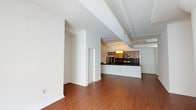 This single-floor unit showcases an industrial-loft aesthetic characterized by prominently exposed cylindrical ductwork running across the elevated ceilings, which contributes to its open, airy atmosphere. The main living area is finished with warm-toned hardwood-style flooring that extends throughout, creating a cohesive and inviting environment. Track lighting along the ceiling complements the industrial design while providing functional illumination across the spacious open floor plan.

The kitchen is thoughtfully designed with warm wood cabinetry, dark countertops, and a textured tile backsplash. Stainless steel appliances, including a refrigerator, microwave, and range, provide modern convenience. A peninsula with a dark countertop doubles as a breakfast bar and separates the kitchen from the living area, making it ideal for both meal preparation and casual dining. A pendant light fixture above the kitchen adds a polished touch.

The bedroom features neutral carpet flooring, high ceilings with continued exposed ductwork, a closet, and a large window fitted with vertical blinds that allows for natural light. The bathroom includes wood-tone cabinetry with a single-sink vanity, a wall-mounted mirror, tile flooring, and a tiled tub-shower combination, offering functional and clean finishes throughout.