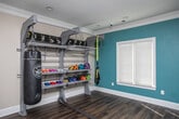 This weight room is thoughtfully designed to support a wide range of fitness activities within a polished, functional environment. Rich hardwood flooring runs throughout the space, complemented by neutral beige walls and a vibrant teal accent wall that adds energy to the room. Crown molding and recessed lighting lend a refined finish rarely found in dedicated workout spaces.

A substantial multi-functional steel rack system lines one wall, organizing a colorful collection of dumbbells, resistance bands, TRX suspension straps, and a hanging heavy punching bag — ideal for strength and combat training alike. A battle rope extends across the floor for conditioning work. An adjustable weight bench sits atop a protective rubber mat near a bank of large windows that flood the space with natural light and offer views of lush greenery outside.

A floor-to-ceiling mirror spans one wall, enhancing the sense of openness and supporting form-checking during workouts. A wall-mounted folding exercise mat and stability balls in blue and yellow provide options for stretching and core training. The space also features a wall-mounted hand sanitizer dispenser, fire extinguisher, and clearly marked exit signage, ensuring safety and convenience throughout.