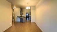 This single-story space showcases an open-concept design that seamlessly connects the living area to the kitchen. Warm-toned wood-look flooring runs throughout, creating a cohesive and inviting atmosphere across the main living spaces.

The kitchen is equipped with gray cabinetry, dark countertops, and black stainless steel appliances, including a full-size refrigerator and microwave. A peninsula with seating overhang serves as both a functional workspace and a casual dining area, effectively defining the boundary between the kitchen and living space.

A decorative multi-light chandelier hangs above, adding a stylish focal point to the open area. The neutral white walls provide a clean backdrop that complements the gray and wood tones throughout the space. A hallway visible in the background suggests additional rooms beyond the main living area.

The layout is practical and efficient, with the kitchen positioned to allow interaction with the living area, making it well-suited for both everyday living and entertaining. The overall design balances modern finishes with functional elements.