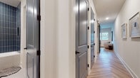 This single-floor residence showcases a cohesive contemporary design with carefully selected finishes throughout. The hallway sets an immediate tone with its sleek gray panel doors trimmed in white and fitted with matte black hardware, leading seamlessly through the home over warm wood-look flooring. Natural light filters in from a window at the end of the corridor, creating an inviting sense of depth.

The kitchen is a standout space, featuring light mint-green flat-front cabinetry with black hardware, complemented by dark stone countertops and a striking blue-gray tile backsplash. Under-cabinet lighting illuminates the workspace, while integrated appliances — including a wall oven, refrigerator, and dishwasher — provide a streamlined, functional layout. Open shelving adds a touch of character with space for greenery and décor.

The bathroom is clean and modern, with large-format white floor tiles and a bold deep-navy vertical tile surround in the tub-shower combination. White cabinetry with black hardware maintains the home's consistent design language.

The bedroom is a serene retreat, featuring warm wood-look flooring, soft neutral walls, and a generous walk-in closet with built-in shelving. A high transom window allows additional natural light to filter in, enhancing the room's comfortable and restful ambiance.