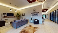 The Whippany Village Apartments community space is a thoughtfully designed, single-floor facility that blends modern sophistication with industrial-chic character. Guests enter through a sleek glass-paneled vestibule featuring marble-style tile flooring, which continues into the main lobby. The branded reception area commands attention with bold Whippany Village signage mounted on a neutral accent wall, complemented by coffered ceiling details with ambient LED cove lighting, a sculptural antler chandelier, plush seating, and a cowhide area rug that adds warmth and texture.

Adjacent lounges offer varied gathering spaces, including a sunlit room with soaring ceilings, rich hardwood flooring, a dramatic exposed brick fireplace wall, a wall-mounted television, and a large window overlooking the outdoors. A dedicated kitchenette and dining nook features the same brick accent wall, blue cabinetry, open wood shelving, and a communal high-top table with industrial-style seating. A separate game room provides entertainment with a foosball table, flat-screen TV, and stylish lounge seating under linear pendant lighting. Throughout, curated artwork, lush tropical plants, and cohesive furnishings create an inviting, upscale communal environment.
