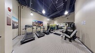 Located at 4720 Rosedale Ave in Bethesda, Maryland, this fitness center offers a well-organized, single-floor workout facility designed to accommodate a comprehensive range of exercise needs. The space features notably high ceilings with an exposed dark ceiling structure, lending an industrial aesthetic, while pendant-style lights provide bright, even illumination throughout the room.

The cardio section is equipped with multiple treadmills and an elliptical trainer, positioned along one side of the space atop protective rubber mats. Opposite this area, a multi-station cable and strength machine — the Instinct series — offers a variety of resistance training options. A dedicated free weights section includes a full dumbbell rack, medicine balls, and a colorful assortment of stability and exercise balls organized on racks along the wall. Large floor-to-ceiling mirrors expand the visual depth of the room and assist users with form and technique.

The flooring consists of light-toned, striped fitness flooring that is both durable and easy to maintain. Wall-mounted large-format photographic prints of the Bethesda cityscape add a decorative touch. Practical amenities include a water cooler station, hand sanitizer dispensers, a waste receptacle, and safety signage. The space is accessed through a standard commercial door with an illuminated exit sign, ensuring code compliance and easy egress.