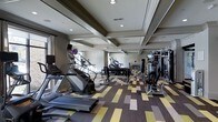 This fitness center is a thoughtfully designed workout space that combines functional equipment with polished architectural details. The room features a coffered ceiling with recessed lighting, lending the space a refined, upscale feel while providing ample illumination throughout. Large mirrors line the walls, visually expanding the area and allowing exercisers to monitor their form during workouts.

The cardio zone is well-represented, with Matrix-brand treadmills and stair climbers positioned toward the front of the room. Moving deeper into the space, a dedicated free weights area includes a full dumbbell rack with clearly organized weights, offering a range of options for strength training. On the opposite side, a multi-station cable and resistance machine provides additional options for full-body workouts.

The flooring features a bold, geometric-patterned carpet in shades of yellow, tan, brown, and gray, adding a contemporary visual element that contrasts with the neutral tones of the walls and ceiling. Natural light filters in through windows along one wall, complementing the recessed lighting. Overall, the space offers a comprehensive and comfortable environment for a variety of fitness routines.