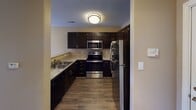 Located at 11206–11222 N Tryon St in Charlotte, NC, this single-floor unit offers a practical and comfortable layout designed for everyday living. The kitchen is a standout feature, outfitted with dark espresso cabinetry, granite-style countertops, and a full suite of stainless steel appliances including a refrigerator, electric range, and built-in microwave. Wood-look flooring runs throughout the kitchen, lending warmth and durability to the space. The open living and dining areas are generously sized, finished with plush wall-to-wall carpeting and neutral warm-toned walls that create an inviting atmosphere. Large windows in the living area allow for ample natural light and include a private exterior door providing direct outdoor access. The bedroom continues the carpeted flooring and features a ceiling fan with light fixture, a window with outdoor views, and clean white trim throughout. The bathroom is well-appointed with a dark-cabineted vanity, a tub-shower combination, wood-look flooring, and a wall-mounted mirror with overhead lighting. The overall design is cohesive and functional, with consistent finishes and a neutral color palette that offers a versatile backdrop throughout the unit.