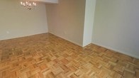 This single-floor space is defined by its standout parquet hardwood flooring, which adds warmth and visual interest throughout the room. The flooring is arranged in a classic block pattern, showcasing natural wood tones with subtle grain variations that lend the space a timeless, elegant character. The walls are finished in a neutral off-white tone, providing a clean and adaptable backdrop that complements the warmth of the wood underfoot. A structural column or partial wall divides a portion of the space, subtly delineating areas while maintaining an open feel. An elegant chandelier is visible in one corner, hinting at a dining or gathering area and adding a touch of refinement to the overall aesthetic. The white baseboards offer a crisp contrast against both the walls and flooring, contributing to a well-finished look. The space feels open and uncluttered, allowing its architectural details — particularly the parquet flooring — to take center stage. Overall, the room presents a neutral, polished environment with classic design elements that offer strong potential for customization and personalization.