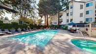 Located at 1277 San Tomas Aquino Road in San Jose, California, this outdoor amenity space centers around a generously sized, crystal-clear swimming pool that serves as the focal point of a well-appointed recreational area. The pool deck is finished with a clean, neutral-toned surface and is surrounded by an array of cushioned chaise lounges and umbrella-shaded seating, ideal for relaxing in the California sunshine.

The space is framed by a rich canopy of mature trees — including towering palms and broad-leafed shade trees — creating a natural, park-like atmosphere that offers privacy and a sense of seclusion. Lush hedging and ornamental plantings line the perimeter, enhancing the green, resort-inspired ambiance.

Dining and gathering areas equipped with tables and chairs provide options for outdoor entertaining, while a decorative fence and structured enclosure ensure a defined and secure environment. The adjacent multi-story building features balconies that overlook the pool area, reinforcing the cohesive relationship between indoor and outdoor living.

Overall, this outdoor space delivers a polished, amenity-rich environment designed for leisure and community enjoyment, making it a standout feature of the property at this San Jose address.