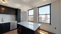 This elegantly appointed high-rise unit combines modern design with refined finishes throughout. The open-concept kitchen serves as a centerpiece, showcasing dark cabinetry with brass hardware, a striking marble-topped center island, a marble tile backsplash, and stainless steel appliances — all set atop rich herringbone-patterned hardwood floors. Large black-framed windows flood the kitchen with natural light while framing dramatic panoramic views of the surrounding city skyline and waterfront.

The living area continues the herringbone hardwood flooring and features a bright, open layout with clean white walls and an entry foyer that provides a welcoming transition into the space. The bedroom offers its own city views through generous black-framed windows, with the same rich hardwood floors and a calm, neutral color palette.

The bathroom is thoughtfully designed with a walk-in glass-enclosed shower clad in white subway tile, a wood-toned vanity with a marble countertop, brass fixtures, and warm stone-look tile flooring. A separate water closet with matching gold hardware accents adds a layer of privacy and convenience. Throughout the unit, a consistent design language of dark, warm, and brass tones creates a cohesive, upscale urban aesthetic.