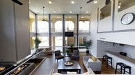 This remarkable space makes an immediate impression with its dramatic double-height ceilings and an expansive wall of floor-to-ceiling windows that flood the interior with natural light. The towering glass panels frame sweeping views of the surrounding outdoor environment, including a spacious patio or balcony area visible beyond, creating a seamless connection between indoors and out.

The main living area is thoughtfully arranged with a generous seating arrangement, featuring neutral-toned sofas and accent chairs accented with yellow throw pillows, centered around a dark wood coffee table. The warm hardwood flooring grounds the space and complements the modern, understated color palette throughout.

Ceiling fans are suspended at height to circulate air through the voluminous room, while recessed lighting provides additional illumination. Dark structural columns punctuate the space, adding architectural definition and visual interest without compromising the open, airy feel.

Lush indoor plants are placed throughout, softening the contemporary aesthetic and adding a natural, organic element. A wall-mounted television and a fireplace are integrated into the design, contributing to the functionality and comfort of the living space. Overall, the space exudes a bright, welcoming ambiance defined by its impressive scale and modern architectural details.