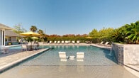 Located at 10480 Auto Mall Pkwy in D'Iberville, Mississippi, this outdoor amenity facility offers a resort-style experience centered around a generously sized swimming pool with crystal-clear water. The pool is a standout feature, equipped with in-water lounge chairs and a cascading waterfall feature along one side, creating a relaxing, spa-like atmosphere. The surrounding pool deck is finished with attractive paver stonework and lined with rows of cushioned chaise lounges and umbrella-shaded seating areas, ideal for sunbathing and socializing under the Mississippi sun. Lush landscaping, including mature palm trees and colorful foliage, frames the perimeter, providing both privacy and a vibrant backdrop. The adjacent clubhouse structure features a hip shingle roof and classic brick accents. A spacious covered patio extends from the building, outfitted with a tongue-and-groove ceiling, ceiling fans, and large dining tables with ample seating — well-suited for gatherings and outdoor entertaining. The facility also includes convenient vending machines and additional seating nooks. The overall design blends Southern architectural charm with modern outdoor leisure amenities, creating an inviting and functional community gathering space.