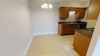 This single-floor space offers a functional and cohesive layout that combines the kitchen and dining areas in an open, approachable design. The kitchen is outfitted with warm medium-toned wood cabinetry, providing ample upper and lower storage, complemented by dark stone-style countertops that add contrast and durability. A tiled backsplash behind the sink area adds a subtle decorative touch, while the under-cabinet lighting enhances both visibility and ambiance. The sink is conveniently positioned along the counter, making the kitchen practical for everyday use. The adjoining dining area flows naturally from the kitchen, creating a seamless transition between the two spaces. Light beige ceramic floor tiles run throughout, lending a clean, neutral foundation that brightens the interior. An elegant chandelier with a classic multi-arm design serves as a focal point overhead, adding a refined touch to the dining space. Neutral light gray walls keep the atmosphere open and versatile, suitable for a variety of furnishing styles. White baseboards provide a crisp finish along the perimeter. Overall, the space presents a tidy, well-maintained environment with a practical and inviting character.