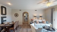 This single-floor residence offers a thoughtfully designed layout with a cohesive neutral color palette and wood-look flooring that flows through the main living areas. The living room serves as the heart of the home, furnished with ample seating, crown molding, a ceiling fan, and a sliding glass door that opens to an outdoor space, allowing natural light to fill the room. An elegant arched entryway connects the living area to the rest of the home, adding a classic architectural detail.

The adjacent dining area features crown molding and direct sightlines to the kitchen, creating an open, connected feel. The kitchen is equipped with white cabinetry, dark countertops, stainless steel appliances including a range, microwave, and dishwasher, along with a double-basin sink.

The primary bedroom offers a cozy atmosphere with plush carpet and a ceiling fan. Additional rooms include a light-filled secondary bedroom with large windows and built-in storage seating, and a versatile flex room currently used as a home office with wall-mounted shelving and a daybed. Both bathrooms are finished with wood-look flooring, white vanity cabinetry, and a tub-shower combination.