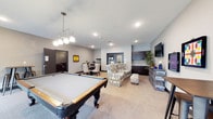 This single-floor Game Room and Theater is a versatile entertainment space designed to accommodate a variety of social activities. The open layout is anchored by a full-size pool table positioned beneath a stylish multi-globe pendant light fixture, creating a natural focal point upon entry. The neutral gray walls and light carpeted flooring provide a clean, contemporary backdrop throughout the space.

A comfortable lounge area is situated alongside the pool table, featuring a patterned accent chair and a plush ottoman, offering a relaxed spot for guests to gather. Nearby, a wall-mounted flat-screen television is positioned above a dark-wood credenza, making this corner ideal for viewing entertainment. Dark-wood upper cabinetry lines the adjacent wet bar area, providing ample storage and serving functionality.

Toward the rear of the space, a dining area with a round table and upholstered chairs offers a more structured setting for casual meals or group activities. Recessed ceiling lighting supplements the pendant fixture, ensuring the entire space is well-lit. A secondary door provides convenient access, contributing to the overall flow and functionality of this well-appointed entertainment area.