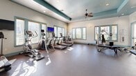 This fitness center is organized across a single level and divided into distinct workout zones, each designed to support a specific type of training. The main exercise floor features rubber flooring and houses a comprehensive selection of strength training machines, including cable systems, leg press equipment, and weight benches. A row of cardio machines — treadmills, ellipticals, and a rowing machine — lines the perimeter, positioned near multiple windows that allow natural light to fill the space and offer views of the surrounding area. A wall-mounted flat-screen television serves this zone for entertainment during workouts.

A separate studio area, finished with warm hardwood flooring, is dedicated to cycling and functional fitness. It accommodates stationary bikes, stability balls in various sizes, medicine balls, and a balance board, making it well-suited for group classes or individual flexibility training. This room also benefits from natural light through its own set of windows.

Throughout the fitness center, tray ceilings with teal accent trim add architectural character, while ceiling fans promote air circulation. Floor-to-ceiling glass partitions and mirrored walls enhance the sense of openness. A large interior window overlooks an adjacent basketball court, further connecting the fitness facility to the broader amenity space.