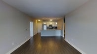 This single-floor space is defined by an open-concept layout that seamlessly connects the living area to the kitchen. The kitchen serves as a functional centerpiece, featuring stainless steel appliances including a refrigerator, microwave, and range, complemented by a dark tile backsplash. White cabinetry provides ample storage, while the granite-topped peninsula offers additional workspace and a natural dividing point between the kitchen and the living area. Three pendant lights hang above the peninsula, adding focused task lighting that enhances the kitchen's contemporary aesthetic. The flooring throughout is a rich, dark hardwood that adds warmth and visual continuity to the space. Neutral-toned walls contribute to a clean, understated palette that keeps the interior feeling bright and cohesive. Along the far wall, a series of built-in storage units and closets with paneled doors provide generous organizational options. The open floor plan creates a spacious, airy feel, making the space well-suited for both everyday living and entertaining. Overall, the design balances modern finishes with practical functionality.
