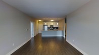 This single-floor space is defined by an open-concept layout that seamlessly connects the living area to the kitchen. The kitchen serves as a functional centerpiece, featuring stainless steel appliances including a refrigerator, microwave, and range, complemented by a dark tile backsplash. White cabinetry provides ample storage, while the granite-topped peninsula offers additional workspace and a natural dividing point between the kitchen and the living area. Three pendant lights hang above the peninsula, adding focused task lighting that enhances the kitchen's contemporary aesthetic. The flooring throughout is a rich, dark hardwood that adds warmth and visual continuity to the space. Neutral-toned walls contribute to a clean, understated palette that keeps the interior feeling bright and cohesive. Along the far wall, a series of built-in storage units and closets with paneled doors provide generous organizational options. The open floor plan creates a spacious, airy feel, making the space well-suited for both everyday living and entertaining. Overall, the design balances modern finishes with practical functionality.