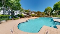 Located at 1575 Ridenour Pky in Kennesaw, Georgia, this outdoor amenity space centers around a generously sized freeform swimming pool with crystal-clear blue water, offering both shallow and deeper sections to accommodate a range of swimmers. The pool deck is finished in a warm tan concrete surface, providing ample space for circulation and relaxation throughout. Surrounding the pool, multiple cushioned lounge chairs and chaise lounges are thoughtfully arranged, inviting residents to unwind and soak up the Georgia sunshine. A covered pergola area adjacent to the pool offers a shaded retreat, furnished with seating and umbrella-topped tables—ideal for casual outdoor gatherings. Stone accent walls and decorative planters add architectural character to the space, while meticulously maintained landscaping featuring lush hedgerows, flowering shrubs, mature trees, and ornamental grasses creates a lush, resort-like atmosphere. Classic black metal fencing defines the perimeter of the pool area, enhancing both safety and aesthetic appeal. Classic lamp posts punctuate the deck, providing ambient lighting for evening use. The backdrop of multi-story residential buildings, partially screened by the mature tree canopy, completes this well-appointed outdoor community space designed for leisure and recreation.